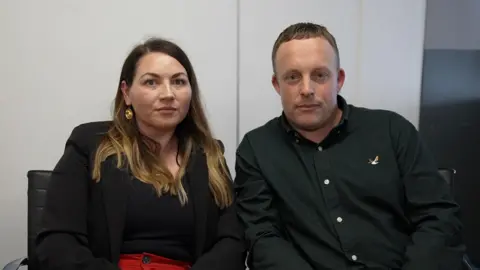 Mark Norman/BBC Liz and Anthony Charlton sit together looking sombre, both are wearing black shirts.