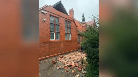 Hawarden High School A pile of broken red bricks are on the floor outside the building, with a hole seen at the side of the roof. There are two large mullioned windows at the side of the building. 