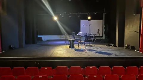 Handout The inside of a theatre, showing the stage. There are red seats at the front. On the stage is a table and chairs. Several lights shine into shot.
