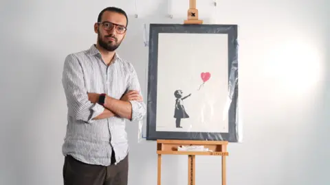 PA Media Gallery manager Lindor Mehmetaj stands with a Banksy painting, Girl With Balloon, at the Grove Gallery in London