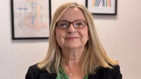 Sam Austin is smiling directly at the camera. She is a white women in her 50's. Sam is wearing rectangular glasses and has shoulder length, blonde hair.
She is wearing a green top with white polkadots on it and a black blazer over her shoulders. 
