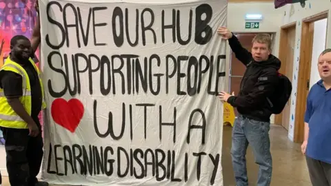Hayley Janceski Two men are holding a large banner indoors, and a third man is in the far right of the photo. One man is dressed in black wearing a high-vis yellow vest, and the second man posing with the banner is wearing a black coat and blue jeans. The banner reads: "Save our hub supporting people with a learning disability".