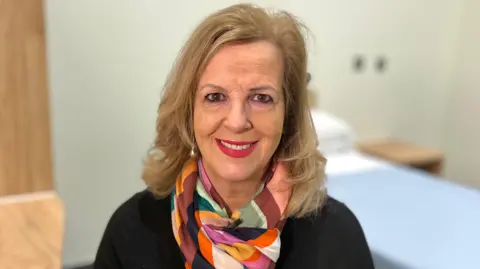 Martin Giles/BBC Caroline Dovan, a lady with blonde hair who is sitting down. She is looking directly at the camera and smiling and she is wearing a black T-shirt, with a coloured patterned scarf. 