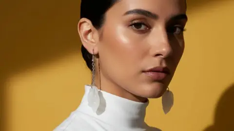 Trash Panda A woman with dark hair standing by a yellow backdrop and wearing a white turtle-neck top. She is wearing long earrings that are white flowers and silver chains.