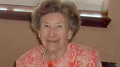 PA Media Una Crown with short curly light brown hair and wearing gold earrings and a pearl necklace and a coral and light pink patterned top. She is smiling and sitting down.