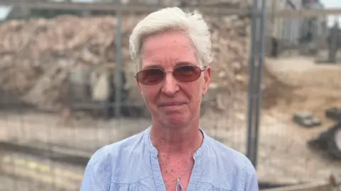 A woman with short blond hair stands in front of a building sight. She is wearing dark glasses, and a blue blouse.