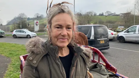 Anette looking at the camera smiling. She is standing at the roadside in front of a relatively small cart and you can see the back of a horse just behind her.