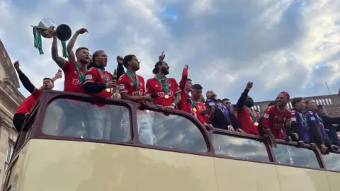 Oli Constable/BBC York City players on an open top bus, with a grand building behind them.