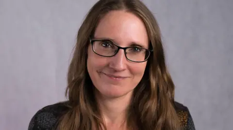 Cambridge Children's Hospital Professor Tamsin Ford is smiling at the camera against a light grey background. She has long brown hair, black square framed glasses and light blue eyes. 