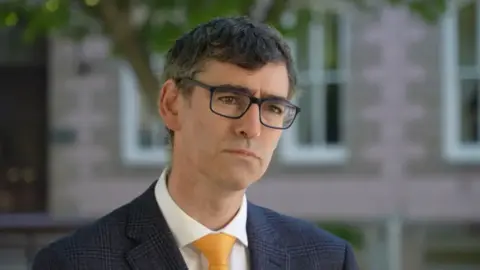 Deputy Montfort Tadier - a man with dark brown hair and a furrowed brow, wearing black rectangle glasses and a navy plaid suit, white shirt underneath and a yellow tie. The background of a pink building and a tree is blurred.