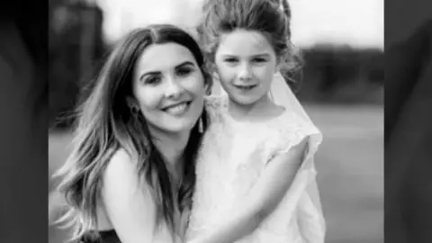 A black and white photograph shows a smiling woman with long dark hair kneeling down beside a smiling young girl who has her hair tied up and is wearing a white dress.