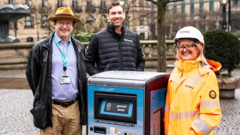 Two men and a woman stand beside a smart waste bin in Sheffield city centre. The man on the left has dark brown hair and glasses, and wears a brown fedora hat, black leather jacket, purple shirt and beige chinos. The man in the middle has short brown hair and a beard, and wears a black puffer gilet and black long sleeved top. The woman has blonde hair and glasses, and wears a white hard hat and fluorescent orange jacket. 