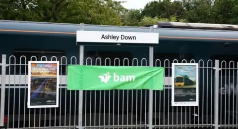 Ashley Down train station sign with Great Western Train in background. The sign is near white railings.