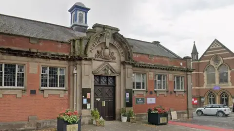 Google A Victorian single-storey red brick library building, with Public Library and a crest carved in stone above a doorway arch. The double doors are dark wood and windows line the building. Outside are two boxes filled with flowers and a white car can be seen on a road to the right, travelling past a church.