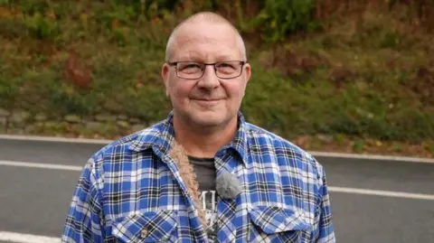 A bald man with glasses wearing a black and blue check jacket. He is stood in front of a road and is looking at the camera with a small smile.