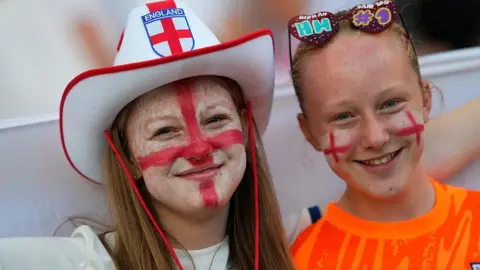 Getty Images A woman and a young girl are pictured wearing an England hat, novelty sunglasses and red and white face paint prior to the UEFA Women's Euro 2025 semi-final match between England and Italy at Stade de Geneve on July 22, 2025