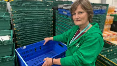 The photo shows a lady with a neutral expression looking at the camera. She is wearing a green hoodie, and is holding an empty blue crate. Behind her are stacks of crates about 5ft (1.5m) high which are also all empty.