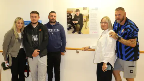 University Hospital Coventry and Warwickshire A family standing next to a wall-mounted artwork in a corridor. There are three people on the left of the picture and two on the right. Naomi Rees-Issitt is pictured in a white shirt. She has blonde hair.