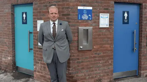 A man with blonde hair stood in front of public toilets.