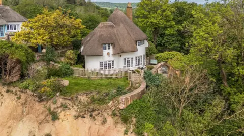 A picture looking from above down at the property and the cliff top. It is a large White House, with a brown roof. There is grassland and trees around the property.
