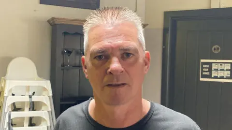 A photo of a white man with grey hair. He is wearing a dark t-shirt. In the background are a stack of white child seats, a glass rack and a brown toilet door