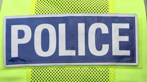 The back of a police officer's florescent yellow jacket, with the word police spelled out in capitals in white, against a blue background