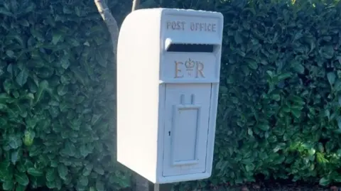 A postbox painted white with gold lettering. Behind it is a hedge.