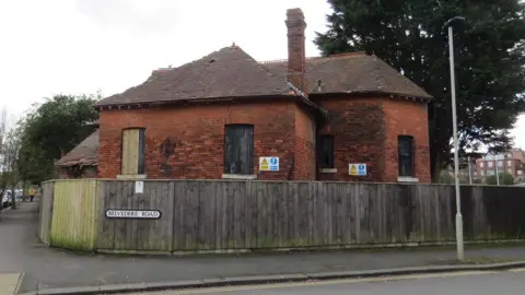 Robert Maw The back of a dilapidated Tudor-style wood and brick building. It has blue and yellow signs on it with safety warnings. It is viewed from the road. A street sign says Belvedere Road.