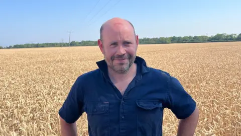 A man with his hands on his hips wearing a denim shirt and he is smiling at the camera.
Crops are behind him 