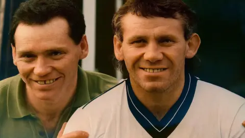 Mike Reid Mike Reid as a younger man, with black hair and a green polo neck t-shirt, smiles at the camera with his hands on the shoulders of Peter Reid, who has lighter hair in a mullet style and is wearing a white England football shirt with a blue v-neck collar. 