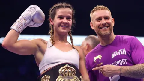 Getty Images Una mujer con chaleco blanco con un guante de boxeo con sonrisas para la cámara: es Terri Harper, campeona mundial femenina. Junto a su toro Steffi, con una camiseta púrpura con logotipos de patrocinador, puntos en la Sra. Harper.