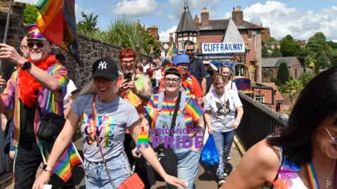 Elliot Benton Crowd walk through the town in rainbow T-shirts and tops carrying rainbow flags.