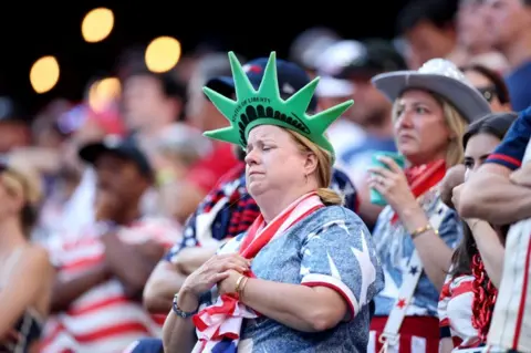 Getty Images A woman in the crowd has her hands clasped together as if praying. She is wearing a green Statue of Liberty crown and a USA scarf. The woman behind her is wearing a cowboy hat.