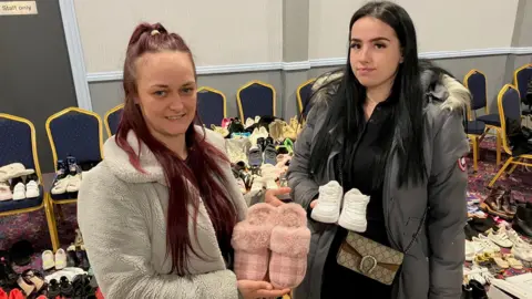 Matt Knight/BBC Rosie Leeman and Jaime-Lee Church standing inside Tilbury Community Centre, each holding a pair of shoes. There are dozens more pairs of shoes behind them.