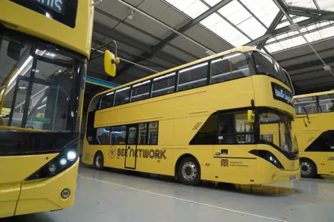 PA Media A row of bright yellow Bee Network buses in a hanger.