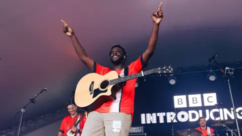 Myles Smith performing at Radio 1's Big Weekend in 2024. He has his arms in the air and is looking out towards the crowd with his guitar round his neck. There is an illuminated BBC Introducing sign behind him. 