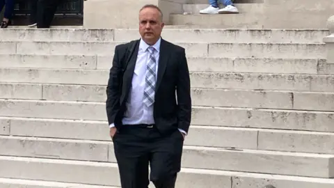 BBC Dylan Dawes walking down the court steps. He's wearing. suit with a chequered tie.