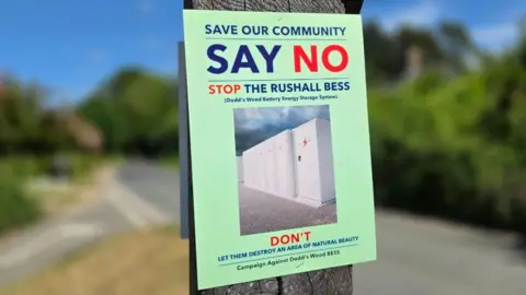 A green sign attached to a wooden post. The sign says "Save our community", "Say no" and "Stop the Rushall BESS".