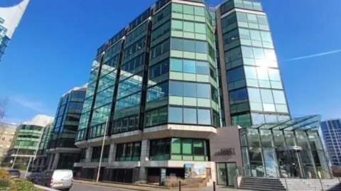 The office complex building, a multi-storey building covered in panes of glass windows which are tinted green. There is a road around the outside of the building, with a silver van parked up on the left. There is a glass entrance, which has a revolving door and a sign which reads Abbey Gardens on the side.
