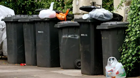 6 bins are lined up on a pavement in front of a house with large bushes. Some of the bins cannot close its lid properly due to excessive carrier bags.