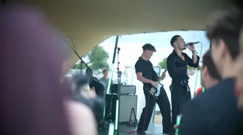 Connor Bennett/ BBC Lead Singer Ben Watts, in all black, singing into his microphone. Meanwhile, Bassist Ed Ruff leans back and strums his guitar.