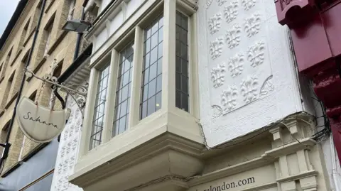 Tom Jackson/BBC A close-up of the first floor of a shop in St Mary's Passage, Cambridge. It has a jutting out window, pained in a stone colour and on either side is white plaster which has fleur-de-lis patterns. 