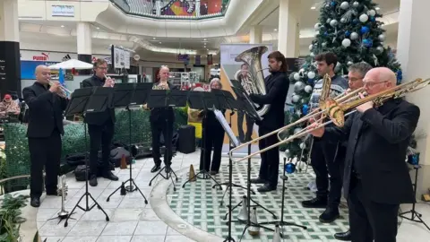 Seven men and one woman play brass instruments in a shopping arcade. They are wearing black suits apart from one man who is wearing a Christmas jumper. They are standing by a Christmas tree that is covered with silver and blue baubles.