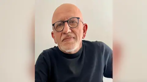 Nicholas Pelas Pelas with a bald head and half grey beard is wearing glasses and a crew neck long sleeved dark shirt. 