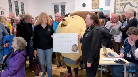 BBC Campaigners in a packed village hall, one of whom is dressed as a hedgehog. Placard says "Hedgehogs live here!"