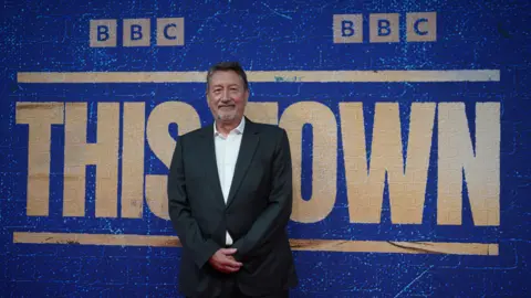 PA Media Steven Knight is wearing a black suit and is standing in front of a large blue backdrop. On the backdrop in big letters it reads "BBC This Town"