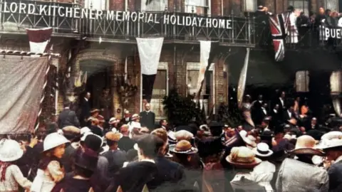 Kitchener's Guest House A colour photo of Lord Kitchener Memorial Holiday Home being opened in 1919. There is a crowd of men, women and children standing in front of the building, which is part of a Victorian terrace. Above the front door is a balcony with letters attached spelling Lord Kitchener Memorial Holiday Home. Flags are flying from its balcony and from the balcony next door.