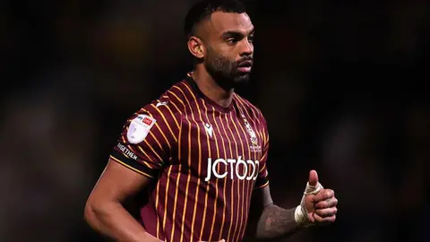 Curtis Tilt in action for Bradford City. He is wearing a maroon and gold striped football shirt with black shorts and socks.