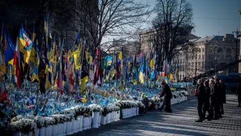 Getty Images A group of people dressed in black watch as a man lays flowers by a collection of flowers and Ukrainian flags. They appear to be in a town square with buildings behind them. It's a sunny day and the sky is blue.