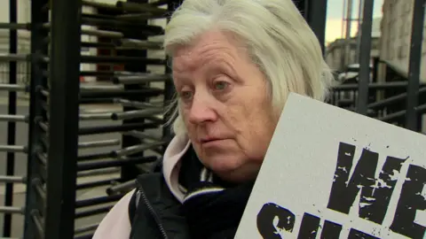 Martina Glenn with cropped grey/blonde hair is standing beside court revolving gates. She has a placard in her hand but only the world We can be seen